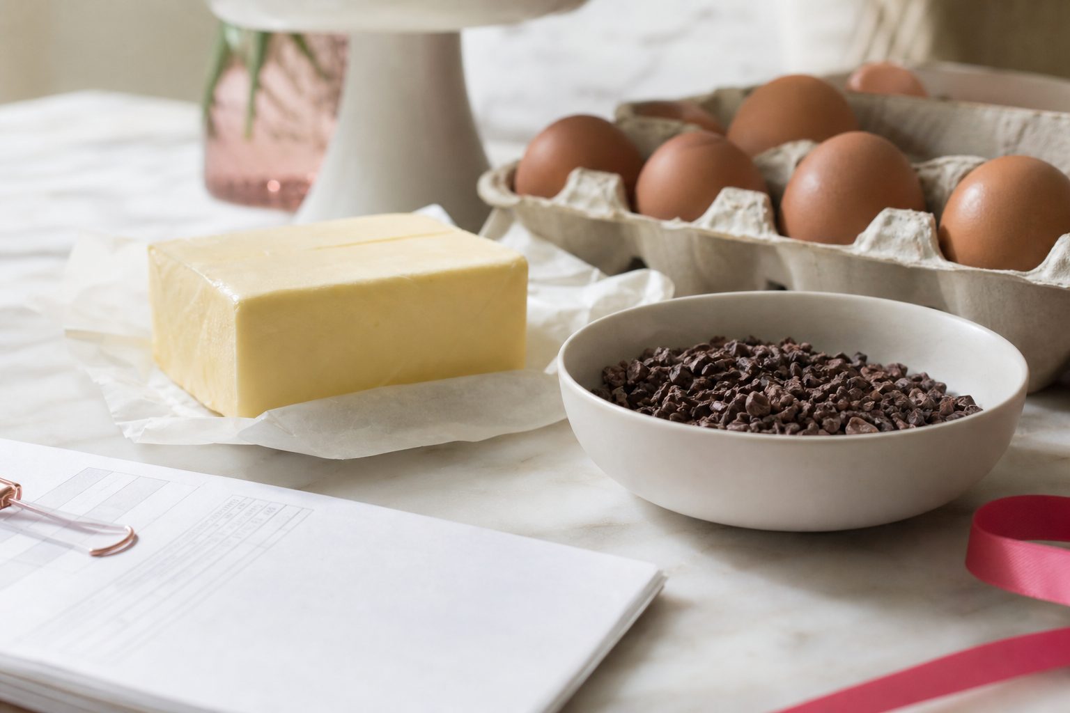 Butter, a carton of brown eggs and a bowl of cocoa nibs beside a folded invoice on a marble counter